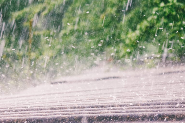 Rain pouring heavily on a wooden deck with blurry green foliage in the background. The image captures the intensity of the downpour with visible raindrops and water splashes on the surface.