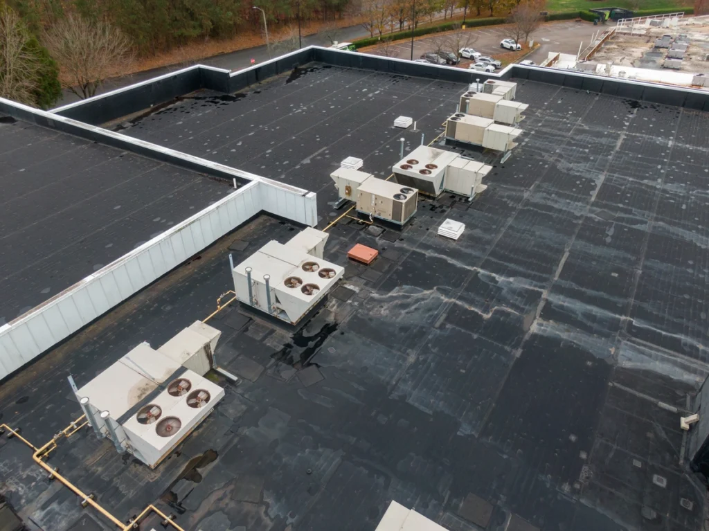 Aerial view of a large flat rooftop with multiple HVAC units. The weathered EPDM roofing shows patches of asphalt and slight water pooling in places. Surrounding the building are trees and a parking area with several cars.