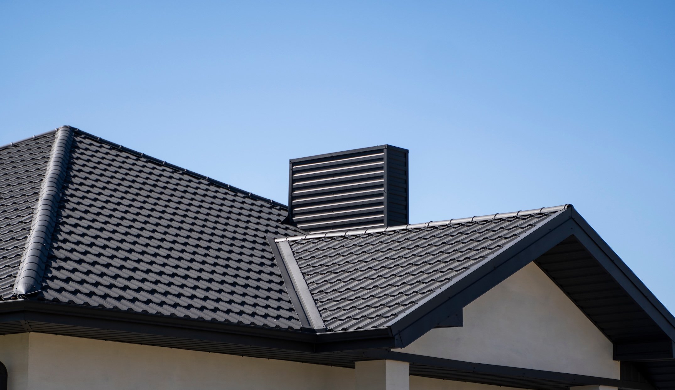 The image shows a close-up view of a modern house roof with dark gray metal shingles and a matching chimney, set against a clear blue sky, showcasing quality metal roofing in Queensbury NY.