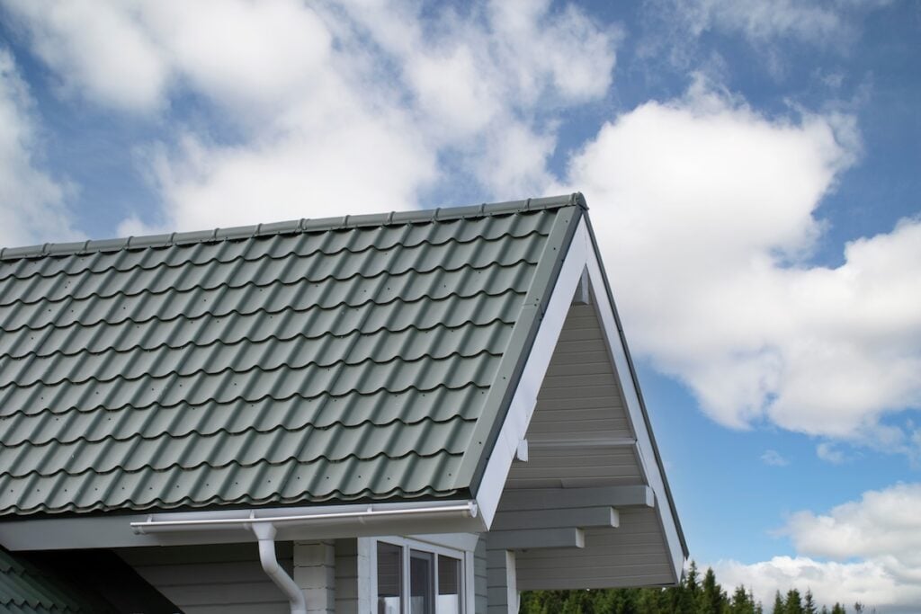A close-up of a modern house roof with green metal tiles, white trim, and guttering showcases stylish metal roofing Ballston Spa NY, set against a partly cloudy blue sky and lush green trees in the background.