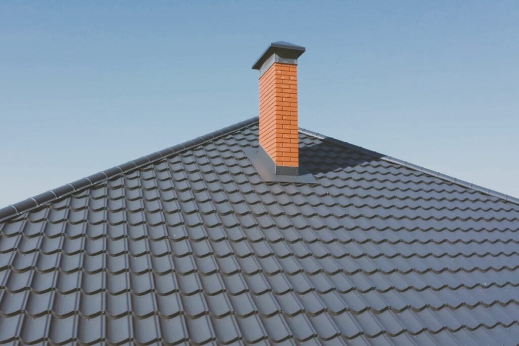 A close-up view of a house roof covered with dark gray metal roofing Queensbury NY, featuring a red brick chimney under a clear blue sky.
