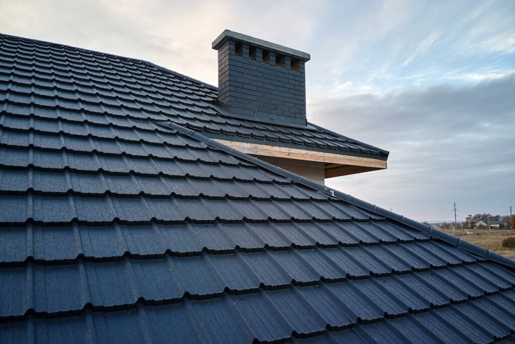 A close-up view of a dark gray metal roofing Queensbury NY installation with a chimney, under a partly cloudy sky. The roof tiles are neatly arranged, and the landscape is visible in the background.