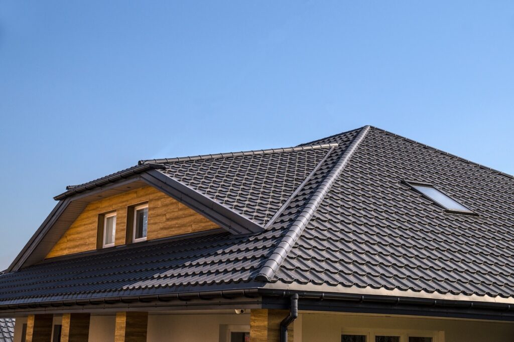 A modern house with a sloped metal roofing in East Greenbush, NY, covered in dark gray tiles and featuring a dormer with two windows and a skylight, set against a clear blue sky.