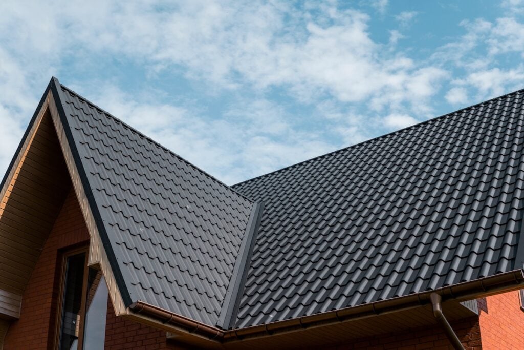 A close-up view of a modern house with a dark gray metal roofing Loudonville NY against a blue sky with scattered clouds. The roof features sharp angles and clean lines, and part of the brick exterior wall is visible.