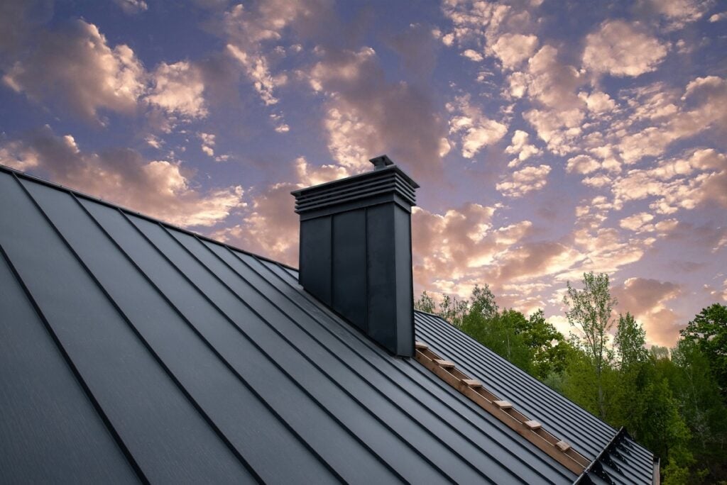 A modern metal roof with a black chimney is shown at sunset, set against a dramatic, cloud-filled sky and green trees—showcasing quality metal roofing East Greenbush NY homeowners can trust.