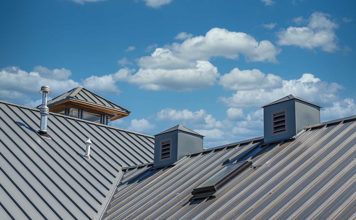 A close-up view of a metal roof with three small rooftop vents, showcasing quality metal roofing in Loudonville, NY, against a bright blue sky dotted with fluffy white clouds.
