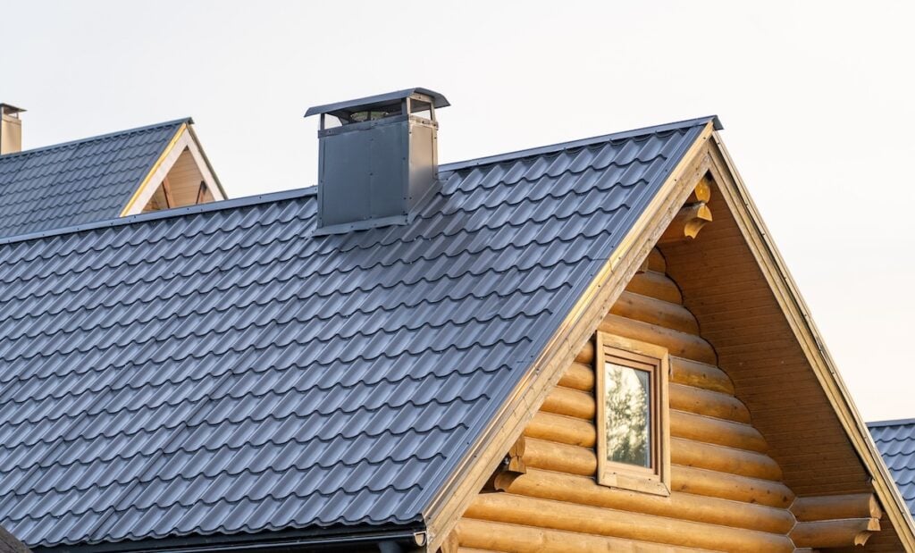 A close-up view of a wooden log house with a gray metal roof, likely installed by metal roofing Loudonville NY experts, featuring a chimney and a window reflecting trees under a clear, light sky.
