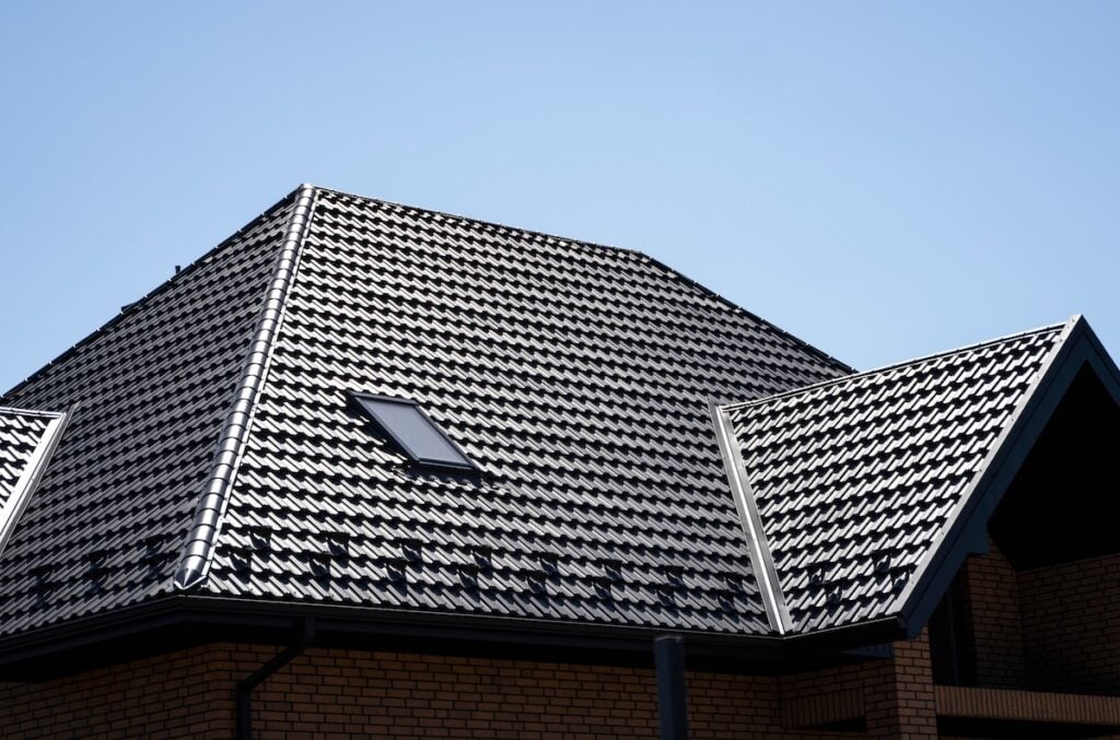 A steeply pitched house roof covered with dark patterned tiles and a skylight window sits beneath a clear blue sky. Light brick walls complete the look, blending classic design with hints of modern metal roofing Hudson NY style.