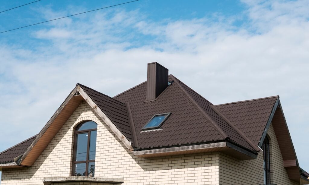 A modern house with beige brick walls and a metal roofing Hudson NY style, featuring dormer and skylight windows, set against a blue sky with scattered clouds.