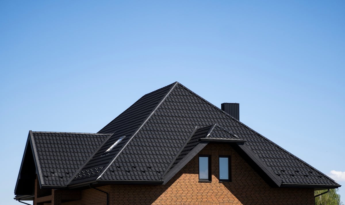 A modern brick house with a dark tiled, multi-gabled roof and several windows, set against a clear blue sky—perfect inspiration for those exploring metal roofing in Hudson, NY.