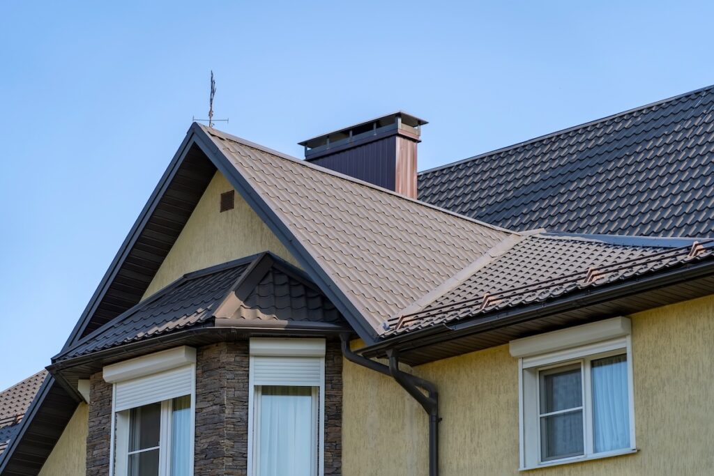 Close-up of a modern house roof with brown metal shingles, installed by metal roofing Averill Park NY, featuring a chimney, beige walls, stone accents, white window frames, and a clear blue sky in the background.