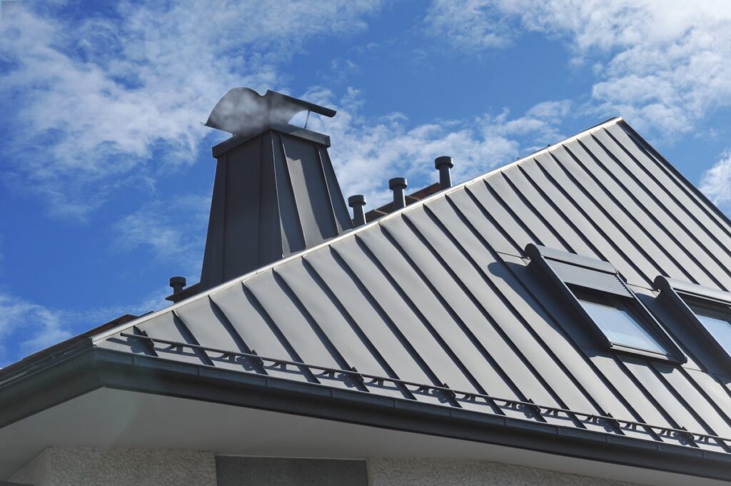 A modern metal roof with vertical seams, multiple vents, a window, and a chimney against a blue sky with scattered clouds showcases the quality of metal roofing Averill Park NY homeowners trust.