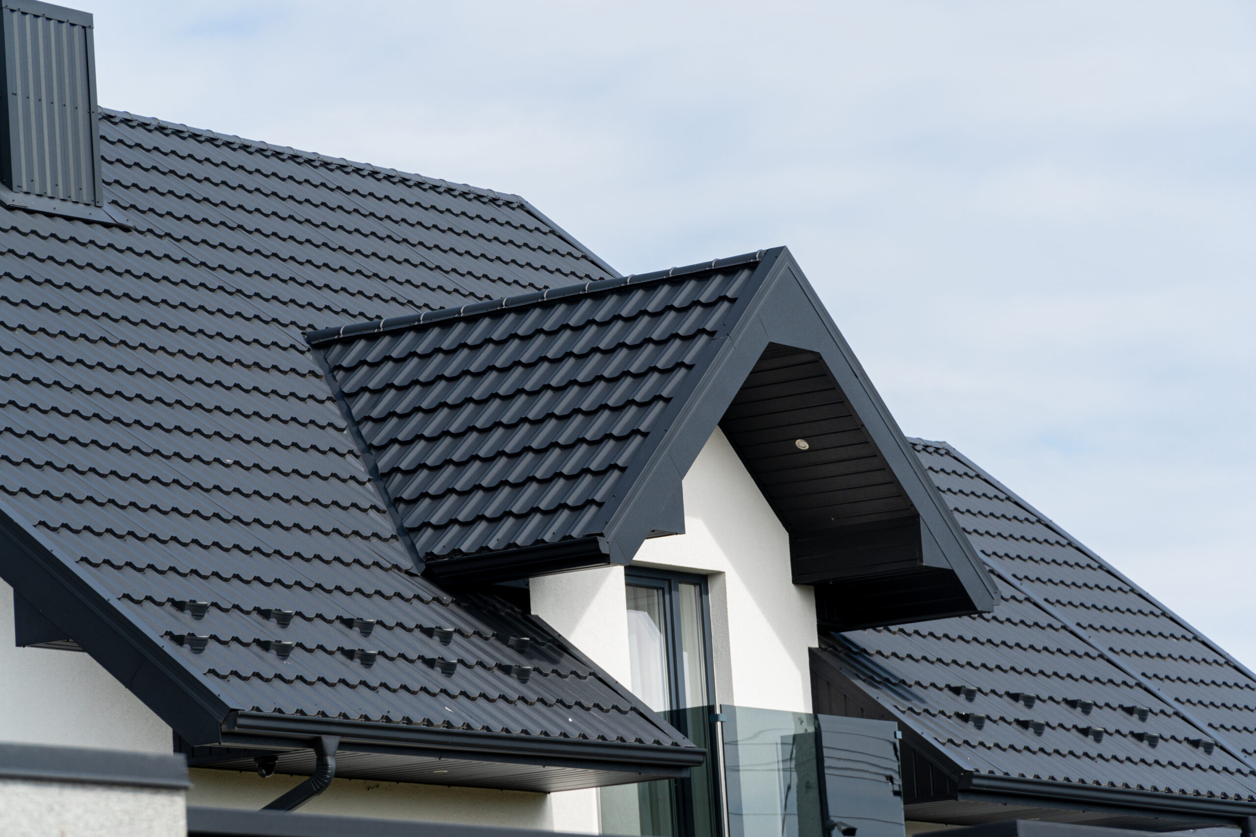 Modern house roof with black metal tiles, featuring a gable and dormer window, showcases quality metal roofing Averill Park NY under a partly cloudy sky.