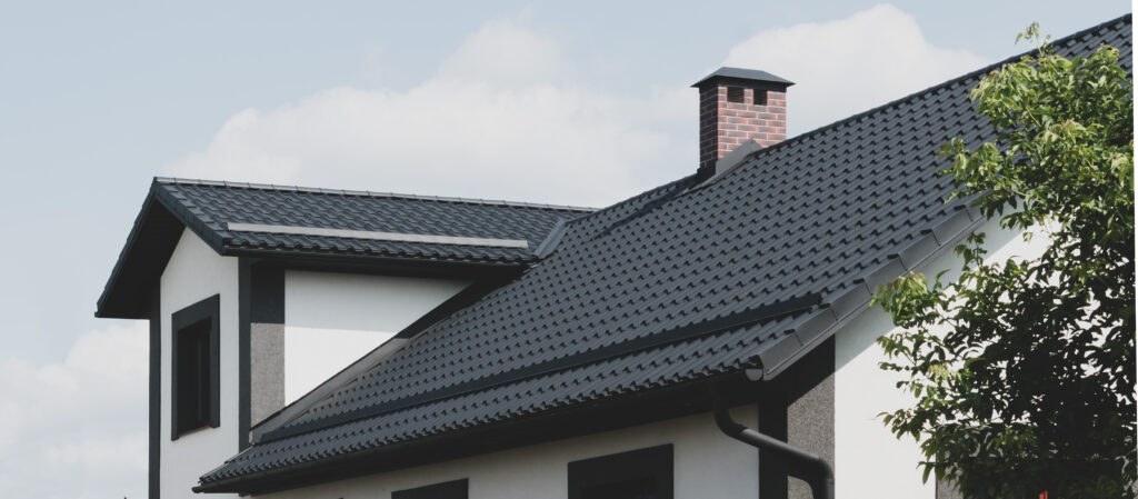 A modern house with white walls, black trim, and a dark metal roofing. A brick chimney rises above the roof, and part of a green tree is visible in the foreground under a partly cloudy Averill Park NY sky.