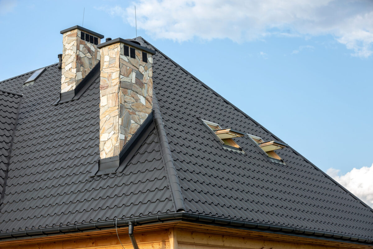 A close-up of a sloped dark grey metal roof with stone chimneys and two skylight windows, showcasing premium metal roofing in Averill Park, NY, set against a blue sky with scattered clouds.