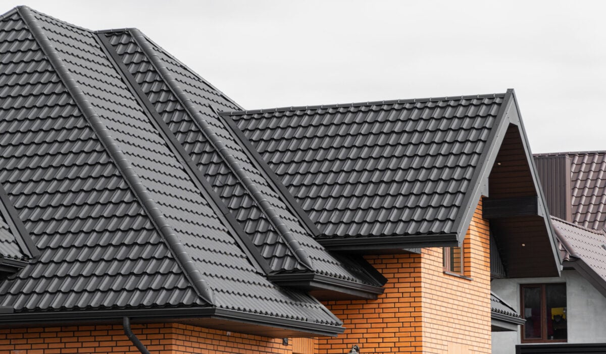Close-up view of a modern house with orange brick walls and a dark gray metal roofing Delmar NY, featuring multiple roof angles and eaves under a cloudy sky.