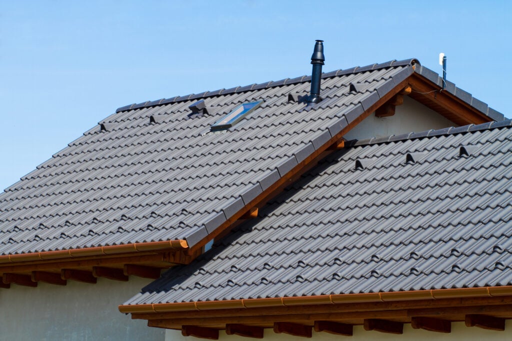Close-up view of a house roof with grey ceramic tiles, a skylight window, multiple small vents, and a chimney—showcasing roofing styles popular in metal roofing Delmar NY—set against a clear blue sky.