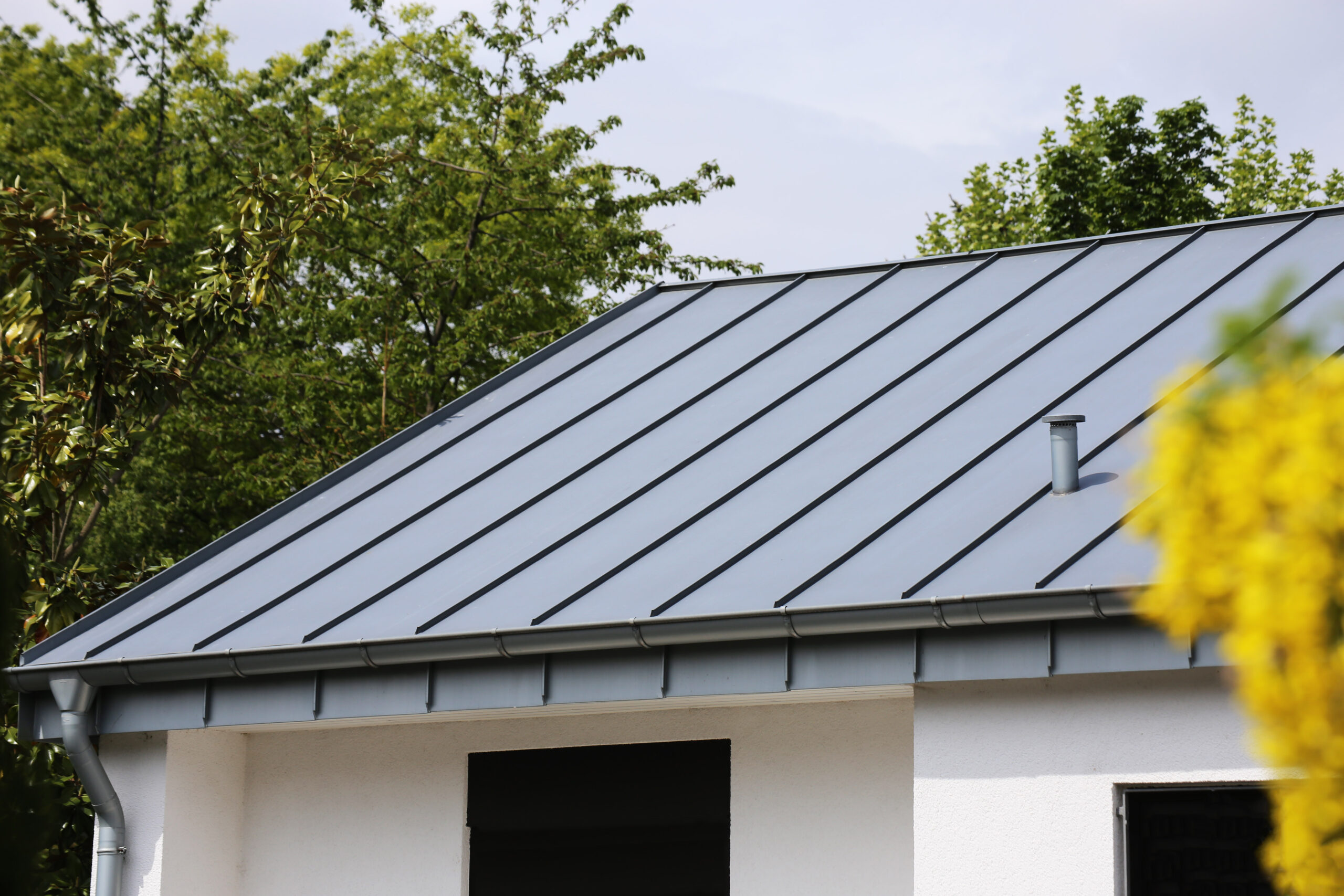 A modern house with a sloped metal roof, light gray in color, surrounded by green trees. The roof features standing seam panels and a small pipe vent—showcasing expert metal roofing in East Greenbush, NY.