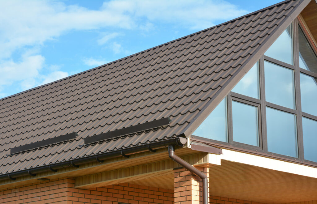 A close-up view of a house with a brown metal tile roof, brick walls, and large windows under a blue sky with some clouds—showcasing quality metal roofing in East Greenbush, NY.