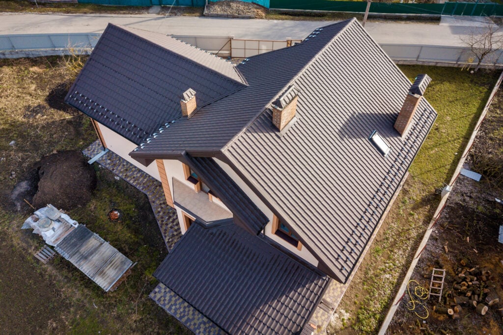 Aerial view of a modern house with a dark gray gabled metal roofing East Greenbush NY, multiple chimneys, and light-colored exterior walls, surrounded by a yard with some construction materials and dirt piles.