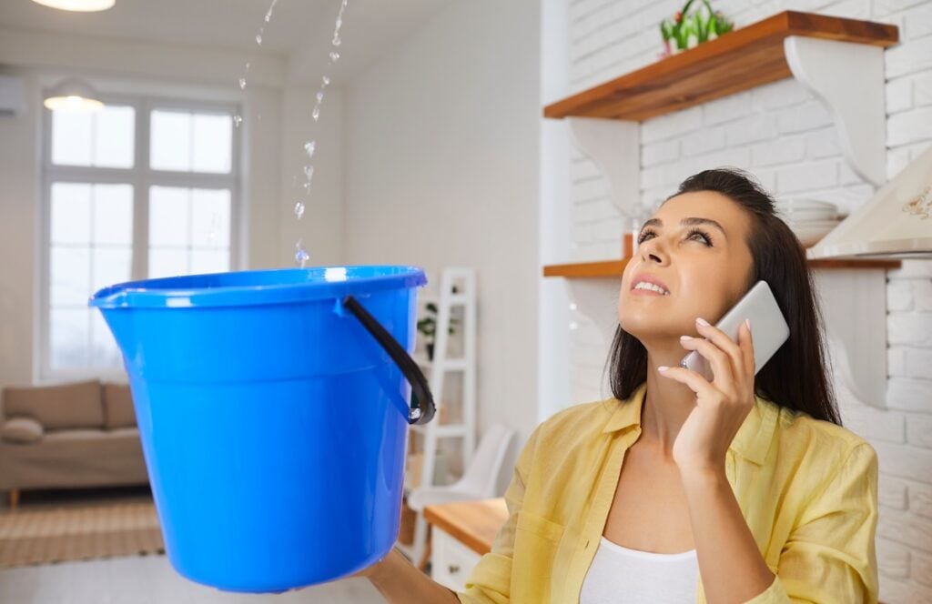 A woman holds a blue bucket under water dripping from the ceiling, likely due to roof problems, while talking on the phone and looking up with concern inside a bright, modern home. roof problems