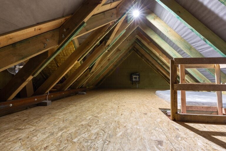 A spacious, unfinished attic with exposed wooden beams and plywood flooring. A bright light shines near the peak of the roof, while some construction materials line the sides—ideal for inspecting or preventing attic rain issues.