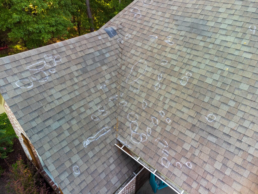 A house roof with numerous chalk circles marking areas of damage or inspection points, seen from above. The roof shingles are light brown, and trees are visible in the background.