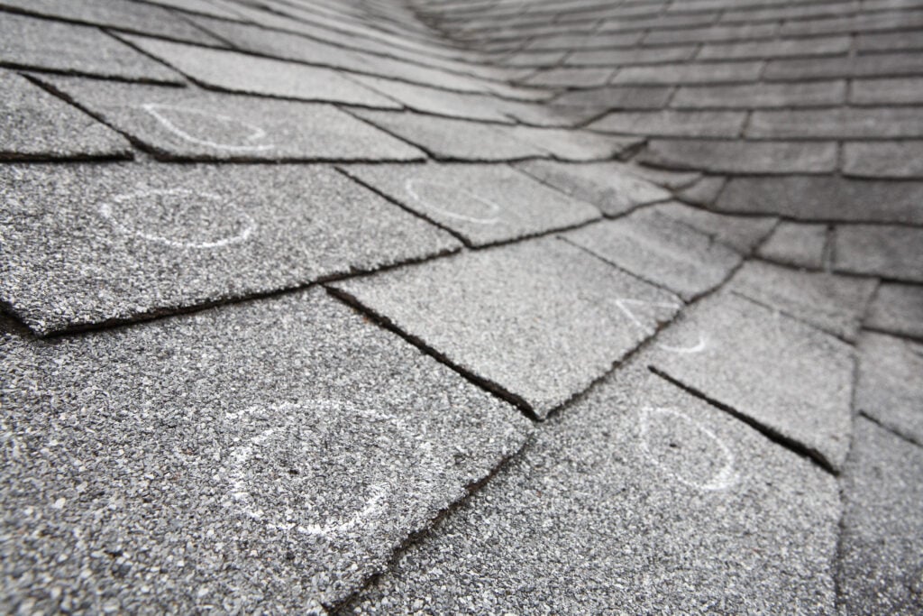 Close-up view of gray asphalt roof shingles with several white chalk circles marking areas of damage or inspection points.