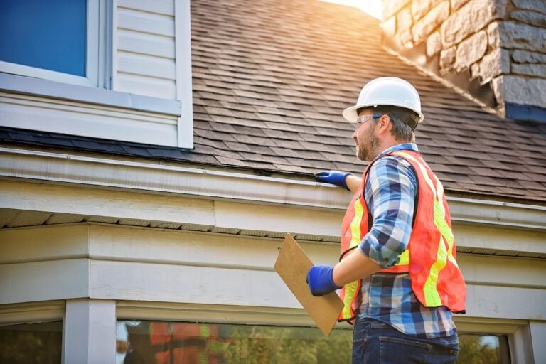 A construction worker wearing a hard hat, safety vest, gloves, and safety glasses inspects the roof and gutter for roof problems, holding a clipboard under one arm. Sunlight shines on the roof and a stone chimney is visible.