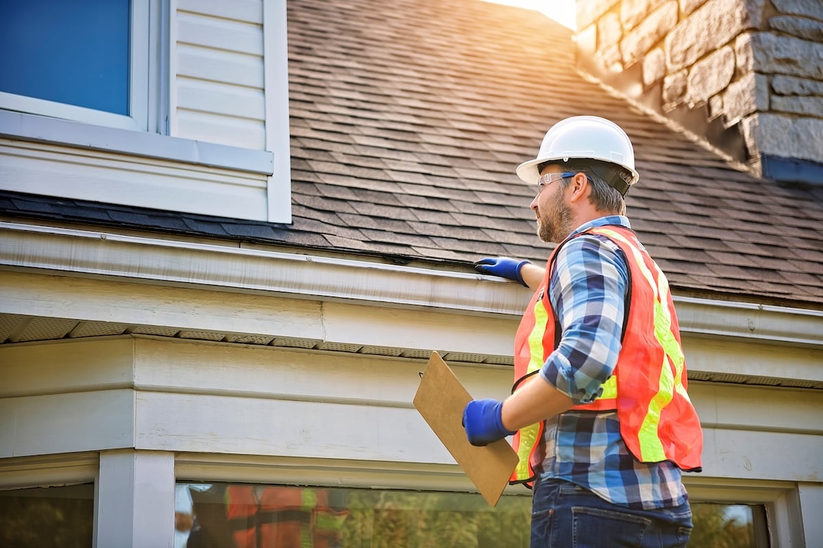 A construction worker wearing a hard hat, safety vest, gloves, and safety glasses inspects the roof and gutter for roof problems, holding a clipboard under one arm. Sunlight shines on the roof and a stone chimney is visible.