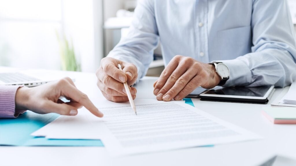 Two people sitting at a desk reviewing and discussing a document; one points at the paper while the other holds a pencil, with a tablet and folders visible on the desk. hail damage vs blistering