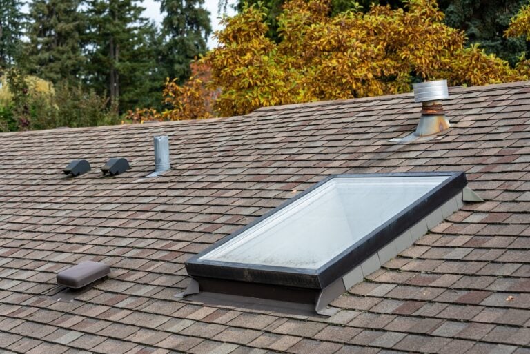 A shingled house roof with a rectangular skylight, several metal vents, and a chimney pipe highlights the need for skylight repair, set against trees with green and autumn-colored leaves in the background.