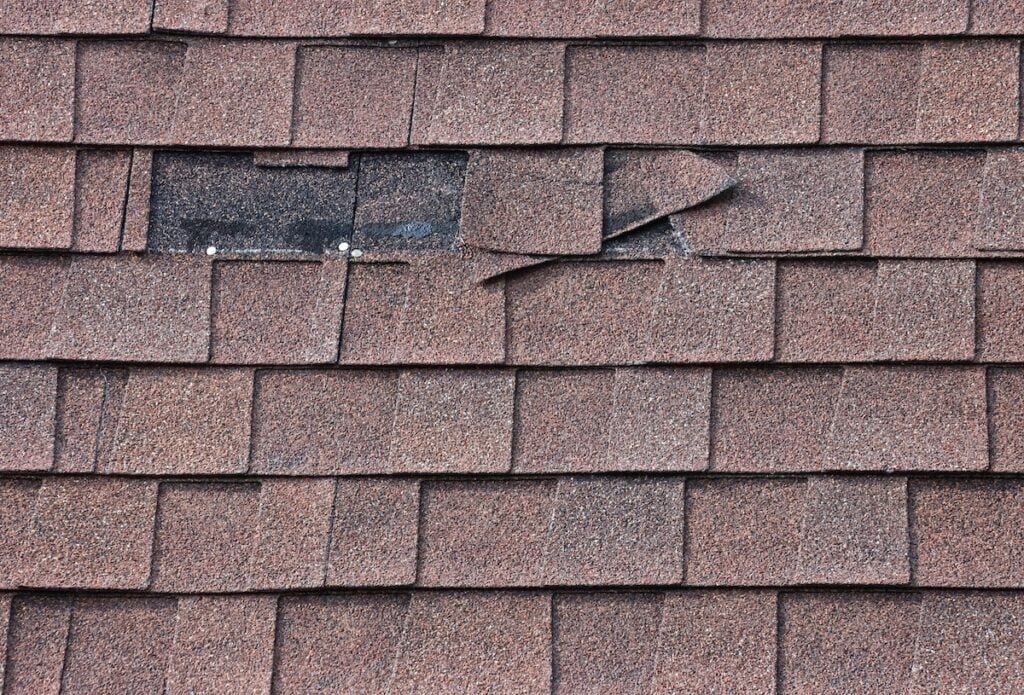 Brown asphalt roof shingles with a few broken and missing shingles indicate roof problems, exposing black underlayment and nails beneath. The damaged area stands out among the otherwise intact shingles.