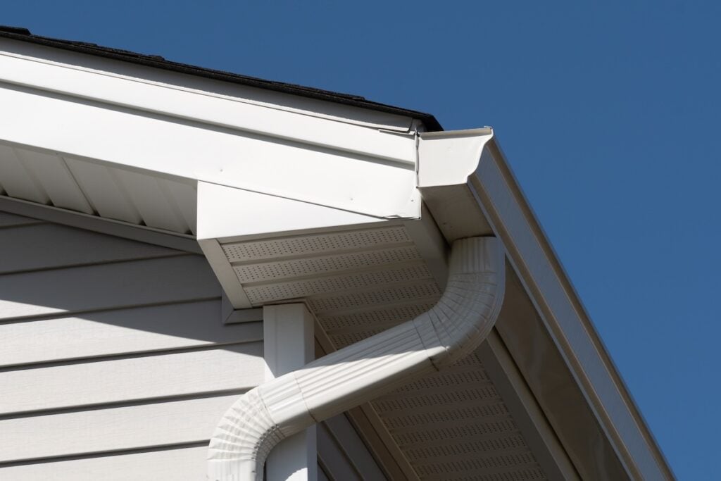 Close-up of a house exterior showing a white gutter system attached to the roof, with a downspout running along the siding, set against a clear blue sky. drip edge