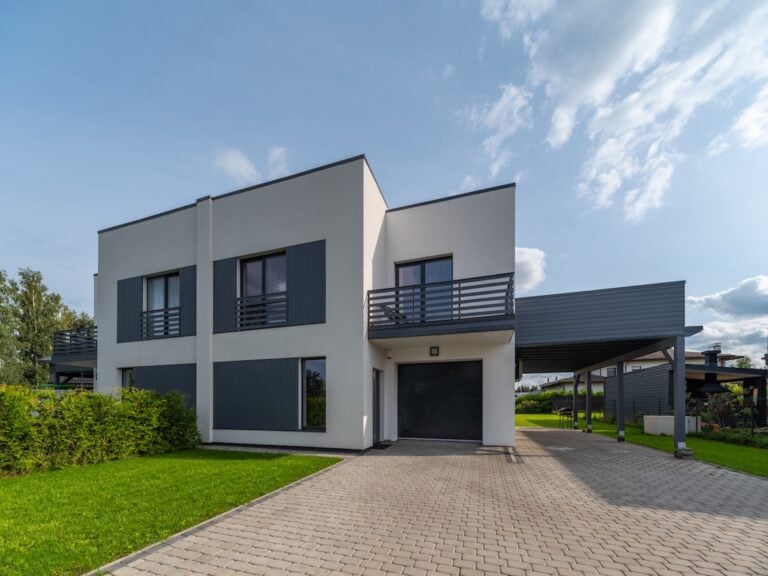 Modern two-story flat roof house with large windows and gray shutters, attached carport, and driveway. The white exterior with gray accents is surrounded by green lawn and shrubs under a partly cloudy sky.