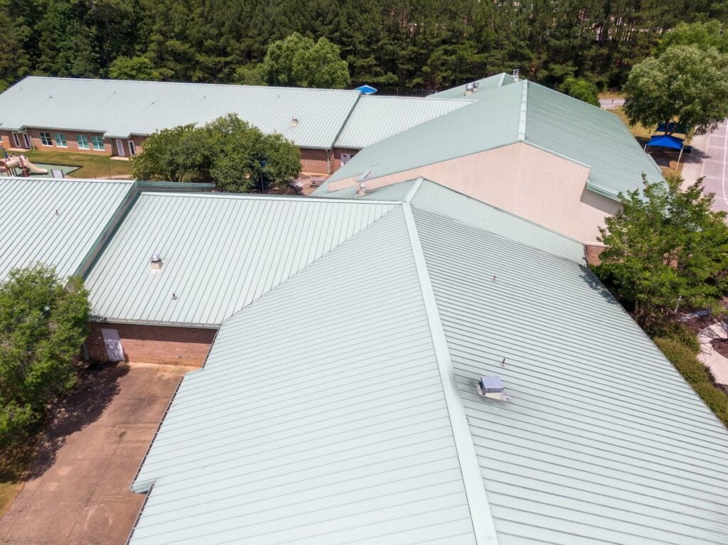 Aerial view of a large building with multiple light green metal roofs surrounded by trees and paved walkways, located in a wooded area. commercial roof types