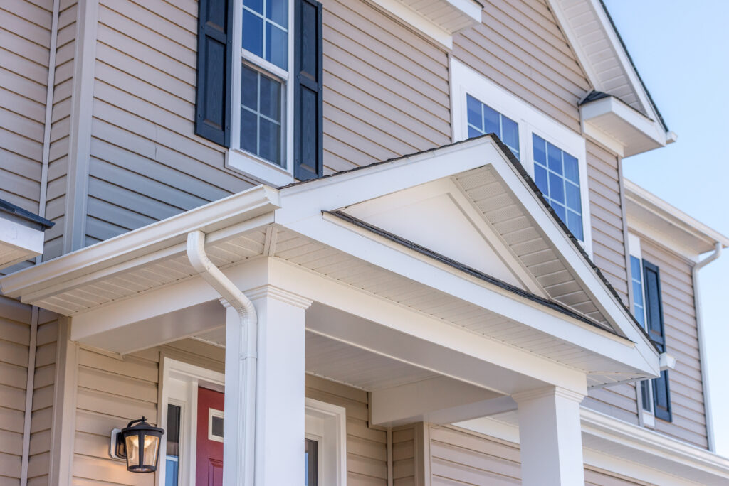 Close-up of a modern house exterior featuring beige siding, white trim, columns, a red front door, black shutters on the windows, and a wall-mounted light beside the entrance. soffit repair