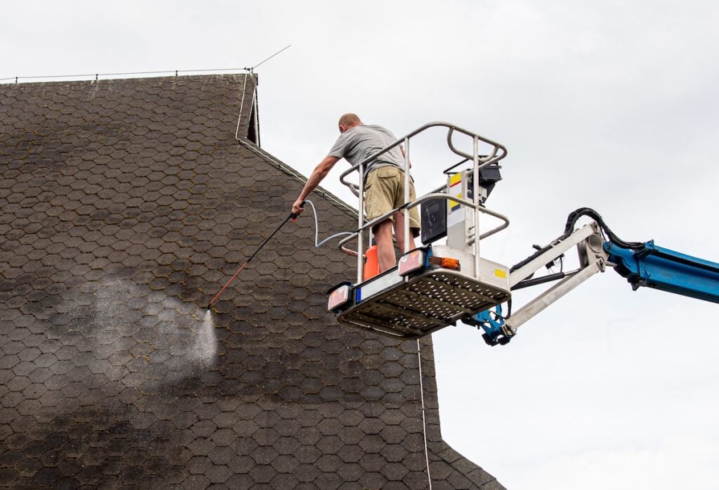 A person standing on a cherry picker uses a pressure washer for roof moss removal, cleaning a large, dark shingle roof under an overcast sky. roof moss removal