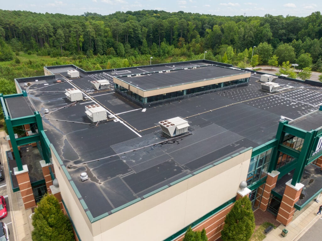 Aerial view of a large commercial building with a flat black roof, multiple HVAC units, and green metal framework, surrounded by trees and greenery. commercial roof types