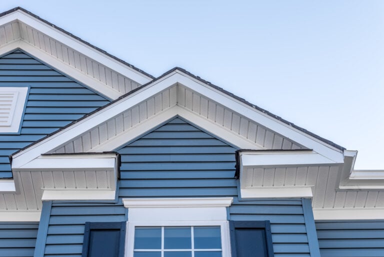 Close-up of the upper exterior of a blue house with white trim, featuring gabled roofs, horizontal siding, a window with white framing, and blue shutters against a clear sky.