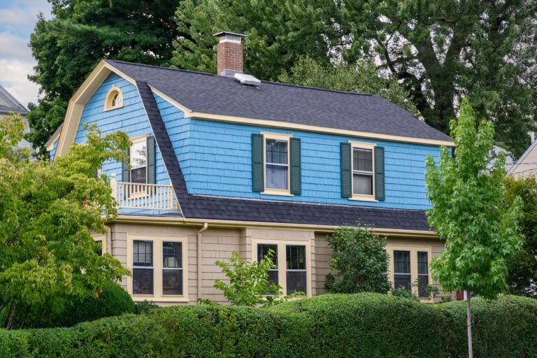 A two-story house with blue siding on the upper level, beige siding on the lower level, green window shutters, a balcony, and surrounded by lush green trees and bushes.