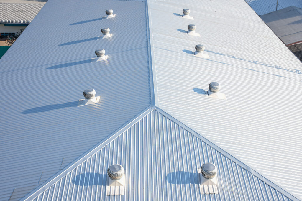 Aerial view of a large corrugated metal roofing with evenly spaced ventilation turbines casting shadows, arranged along the roof’s ridgeline. The roof is clean and bright under sunlight.
