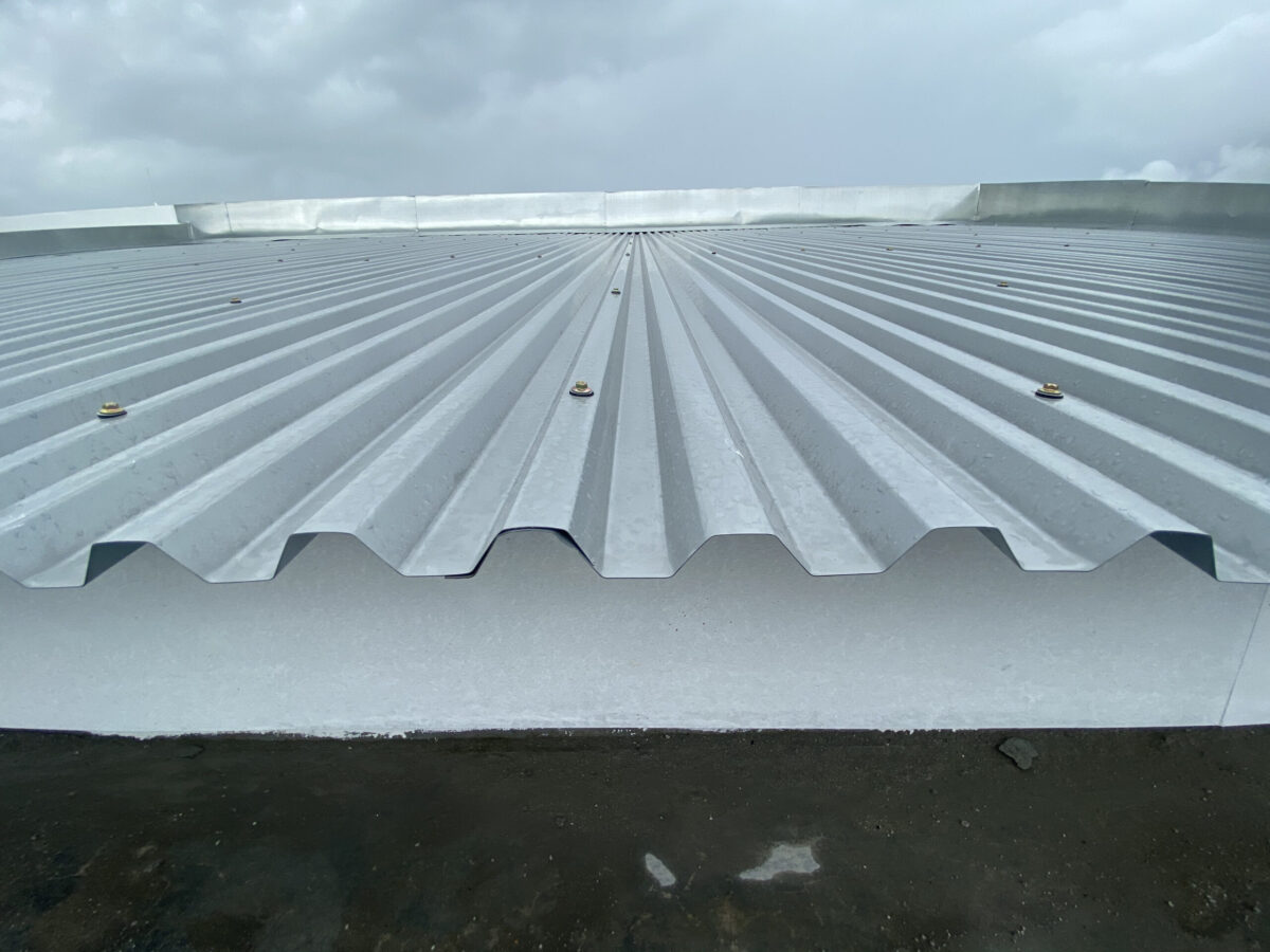 Close-up view of a corrugated metal roofing with evenly spaced screws, set against a cloudy sky. The roof surface appears clean and new.