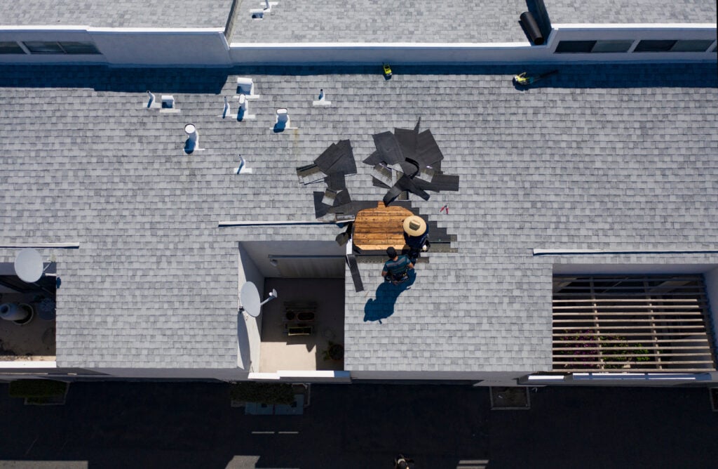 A worker wearing a hat performs shingle roof repair on a gray rooftop, surrounded by tools and roofing materials, as bright sunlight casts shadows across the scene.