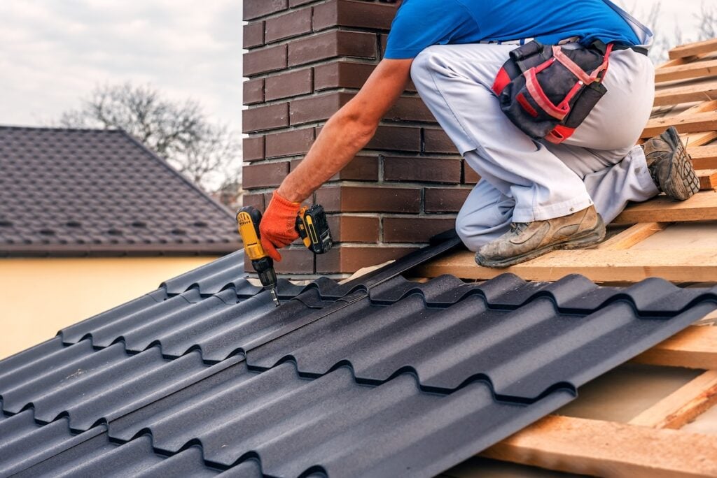 questions to ask a roofer before hiring A worker in gloves and a tool belt uses a power drill to install metal roofing sheets around a brick chimney on a house roof under construction.