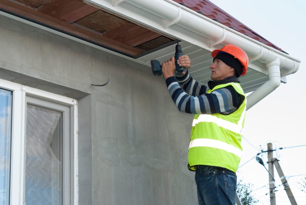 A construction worker in a safety vest and helmet uses a power drill to work on the eaves of a house under a red metal roof on a clear day. questions to ask a roofer before hiring