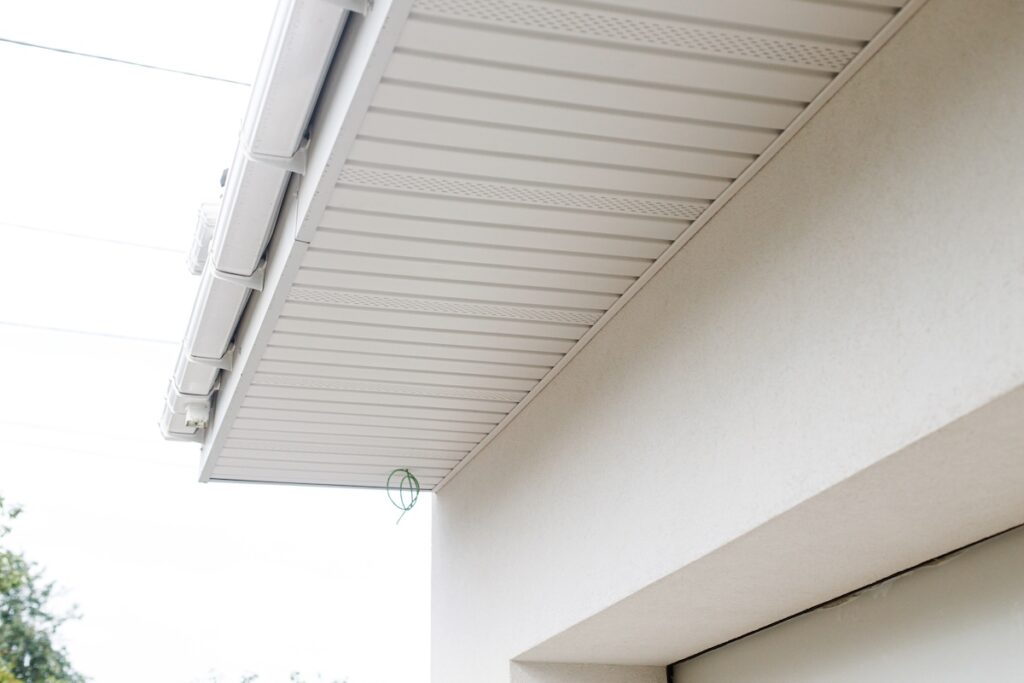 Close-up view of a  soffit repair house’s white soffit and fascia under the roof eaves, with a green twist tie hanging from the edge. The background shows a light sky and some tree branches.