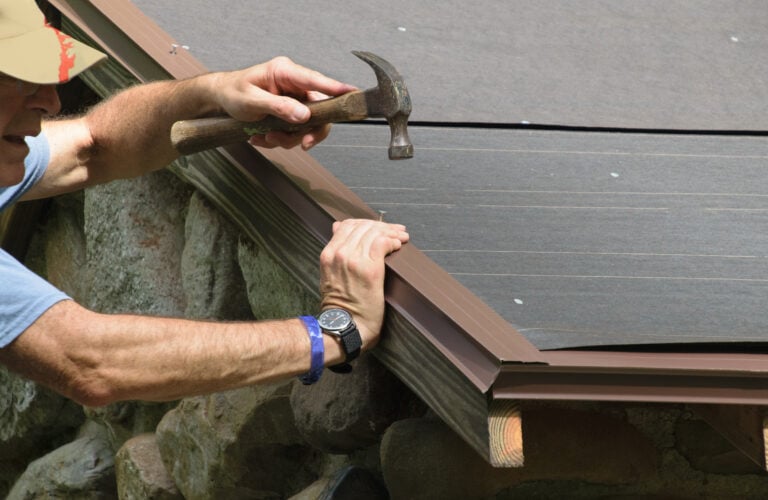 A person wearing a watch uses a hammer to secure the edge of roofing material on a building with stone siding. Only the person’s arms and part of their face are visible. drip edge