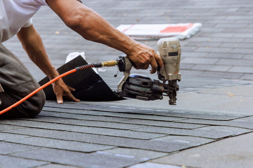 A person installing asphalt shingles on a roof, using a pneumatic nail gun to secure the shingles in place. The photo focuses on their hands and tool, with rows of shingles visible. questions to ask a roofer before hiring