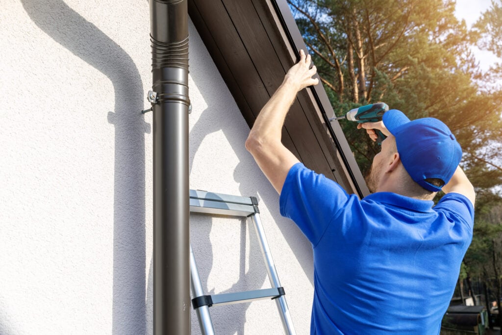 A man in a blue shirt and cap uses a power drill to work on the roof eaves of a house while standing on a ladder, with sunlight and trees in the background.